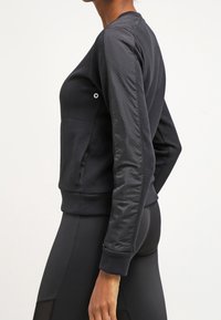 Sudadera negra con un frente liso y mangas texturizadas y perforadas. Presenta un puño y dobladillo acanalados, con un pequeño logo en el pecho.