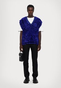 Mėlynas blizgučiais pasiūtas be rankovių liemenė, dėvima ant baltos marškinėlių, derinama su juodomis kelnėmis ir bateliais, rankoje laikoma maža juoda rankinė.