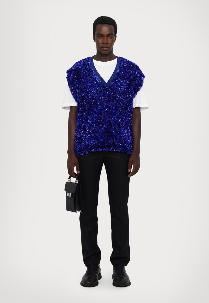 Mėlynas blizgučiais pasiūtas be rankovių liemenė, dėvima ant baltos marškinėlių, derinama su juodomis kelnėmis ir bateliais, rankoje laikoma maža juoda rankinė.