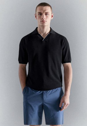 Giovane uomo che indossa una polo nera a maniche corte e pantaloncini blu, in piedi con le mani in tasca davanti a uno sfondo grigio uniforme.