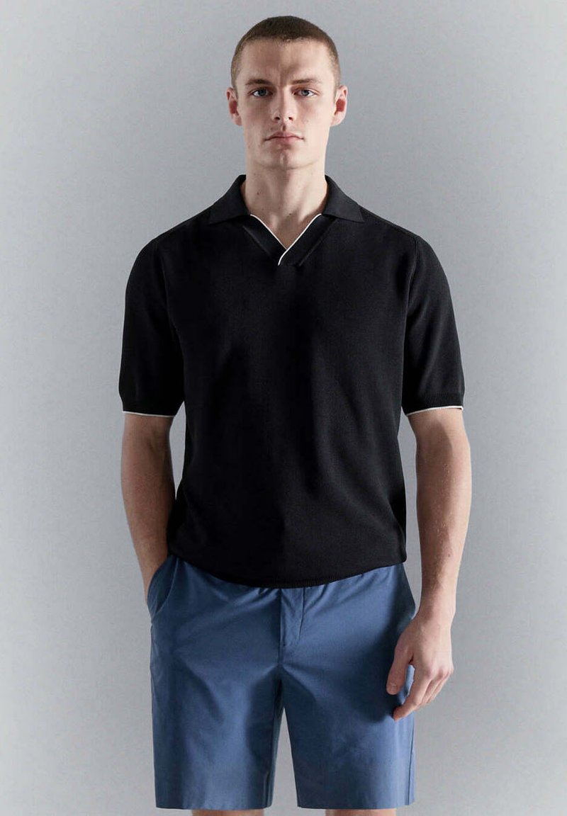 Jeune homme portant un polo noir à manches courtes et un short bleu, debout les mains dans les poches devant un fond gris uni.