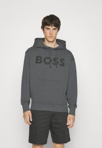 Felpa grigia in cotone con grande logo nero "BOSS", cappuccio con cordino e tasca a marsupio, abbinata a pantaloni corti neri a fantasia.
