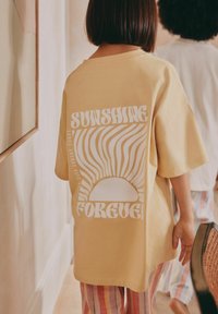 Gelbes, übergroßes T-Shirt mit weißem Sonnenmotiv und dem Text "SUNSHINE FOREVER" auf dem Rücken. Weiches Material, lockere Passform, sichtbare Nähte.