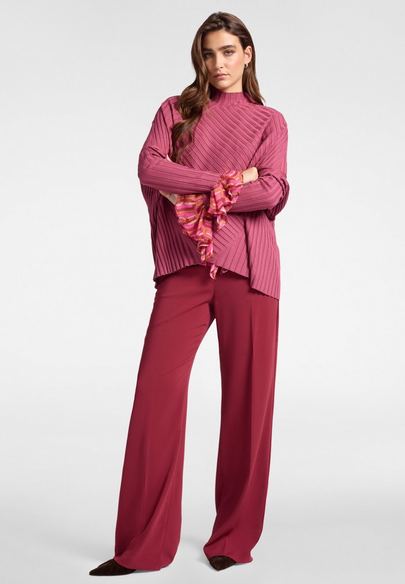 Mujer de pie con los brazos cruzados, vistiendo un suéter rosa acanalado con puños estampados y pantalones anchos burdeos, sobre un fondo sencillo.