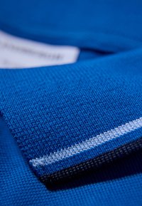 Niebieska dzianina o teksturowej powierzchni. Posiada biały i czarny akcent w paski. Etykieta częściowo widoczna, z instrukcjami pielęgnacji.