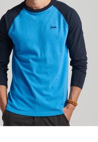 Sininen ja laivastonsininen pitkähihainen raglan-paita, jossa on pyöreä kaula-aukko ja brodeerattu logo rinnassa. Valmistettu pehmeästä kankaasta.