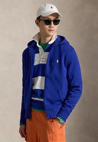 Homme portant une casquette blanche, des lunettes de soleil foncées, un sweat à capuche bleu, un polo rayé bleu et blanc, un pull vert et un pantalon orange sur fond neutre.