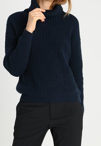 Pull à col roulé en maille bleu marine avec des côtes texturées, manches longues et ourlet ajusté, porté sur un pantalon noir.