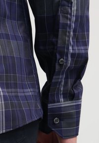 Camisa de cuadros azul marino con puños abotonados, textura suave, líneas más claras en contraste y un ajuste ligeramente entallado en la cintura.