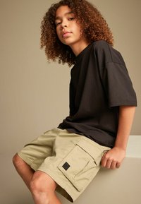 Sort kortærmet T-shirt parret med beige cargo shorts med sidelommer og et tekstureret stof. Modellen sidder opad et neutralt baggrund.
