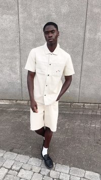 Beige kortermet skjorte med enkel design og to lommer, parret med matchende beige shorts og svarte loafers med hvite sokker.