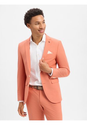 Mężczyzna uśmiecha się, ubrany w koralowy garnitur z białą koszulą i poszetką, poprawiający klapę marynarki na jednolitym białym tle.