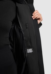 Mână ținând un sacou negru deschis, arătând buzunarul interior cu fermoar și eticheta albă "Khujo original changing threads" pe căptușeală.