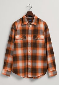 Camicia di flanella a scacchi in arancione, marrone e bianco. Presenta una chiusura con bottoni, due tasche sul petto e un orlo arrotondato.