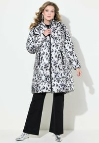 Abrigo acolchado en estampado de leopardo en blanco y negro, con cierre de cremallera, bolsillos laterales, material brillante; combinado con pantalones negros y botines metálicos.