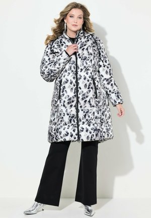 Bunda s kapucí v černobílé leopardí potisku, na zip, s bočními kapsami, lesklý materiál; nošena s černými kalhotami a metalickými kotníkovými botami.