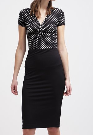 Mujer vestida con una falda lápiz negra y una blusa negra de manga corta con lunares blancos y botones en el escote.