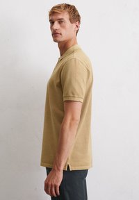 Jasnobrązowa koszulka polo z kołnierzykiem, krótkimi rękawami i bocznymi rozporkami. Wykonana z teksturowanej, oddychającej tkaniny, o prostym wzornictwie.