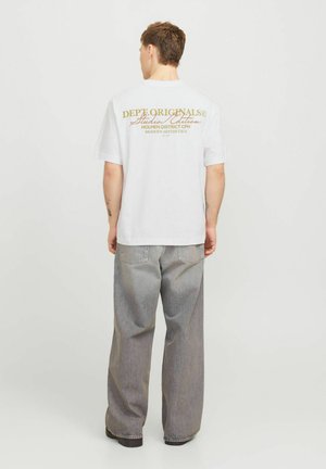 Man draagt een wit T-shirt met gele tekst "Dept Originals Studio Edition" en losse grijze spijkerbroek, staand met de rug naar de kijker.