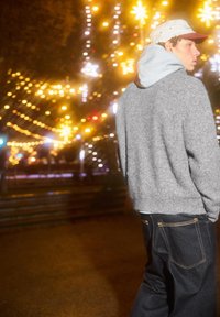 Grau gestrickter Pullover mit Kapuze, getragen über einem hellen Hemd, kombiniert mit dunklen Denim-Jeans. Im Hintergrund verschwommene warme Lichter.