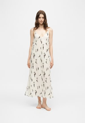 Tânără cu păr lung purtând o rochie maxi albă fără mâneci, cu un model floral negru, stând desculță pe un fundal simplu.