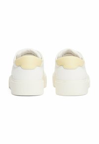 Des baskets blanches avec une semelle en caoutchouc texturé, dotées d'un accent jaune lisse à l'arrière et d'un design arrondi.