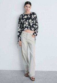Črn cvetlični crop top z dolgimi rokavi in detajli z vrvicami, v kombinaciji s svetlo sivo širokimi hlačami. Obleka ima sproščen kroj.