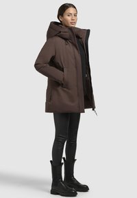 Brown insulated parka with a large hood, side pockets, and a zip closure. Worn over black leggings and ankle boots. Smooth fabric texture.