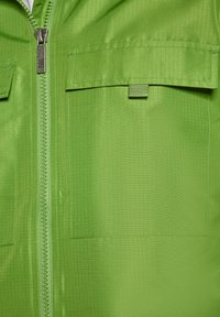 Grön zip-up jacka gjord av slätt, texturerat tyg, med två bröstfickor och en central dragkedja med ett silverfärgat draghandtag.