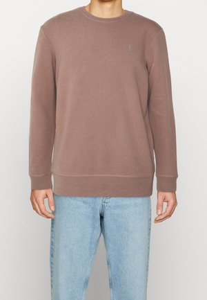 Sudadera marrón con cuello redondo, mangas largas y puños acanalados. Presenta un pequeño logo bordado en el pecho. Se lleva con jeans de color azul claro.