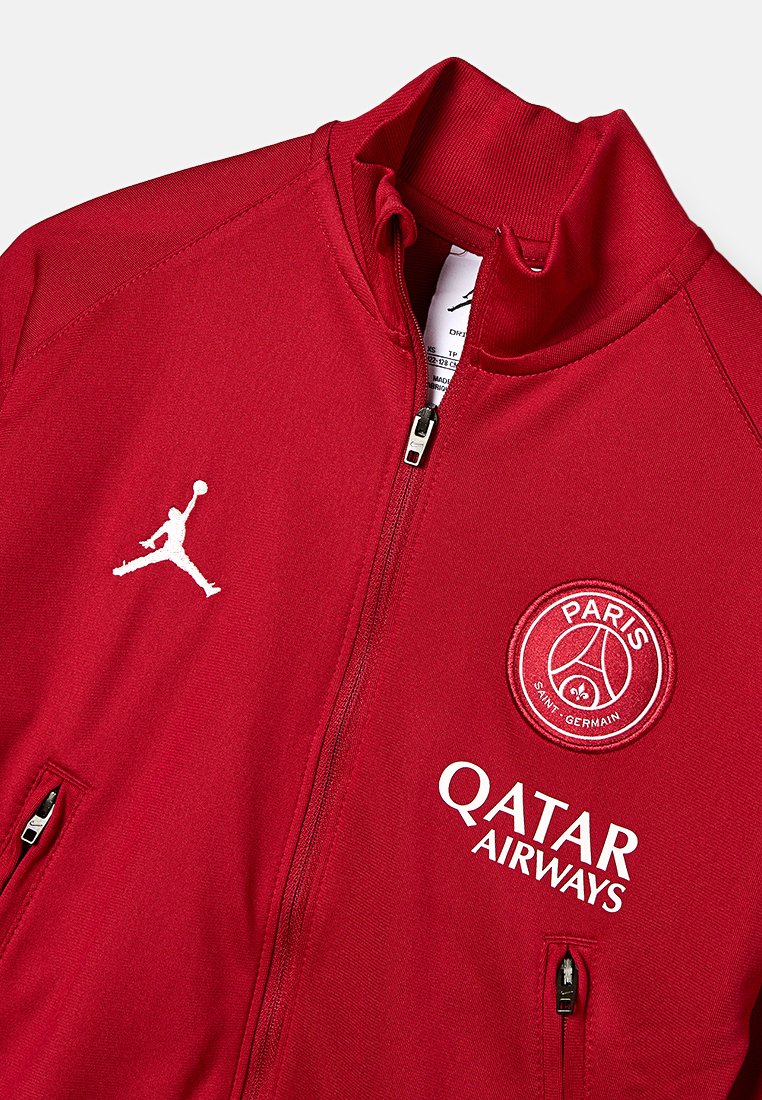 Veste zippée rouge fabriquée en matériau lisse. Affiche des logos brodés en blanc : Jordan, Paris Saint-Germain et Qatar Airways. Dispose de poches latérales.
