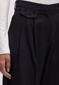 Zwarte op maat gemaakte broek met plooien, met een uniek zakflapontwerp en knoopsluiting. Tentoongesteld naast een witte geribbelde top.