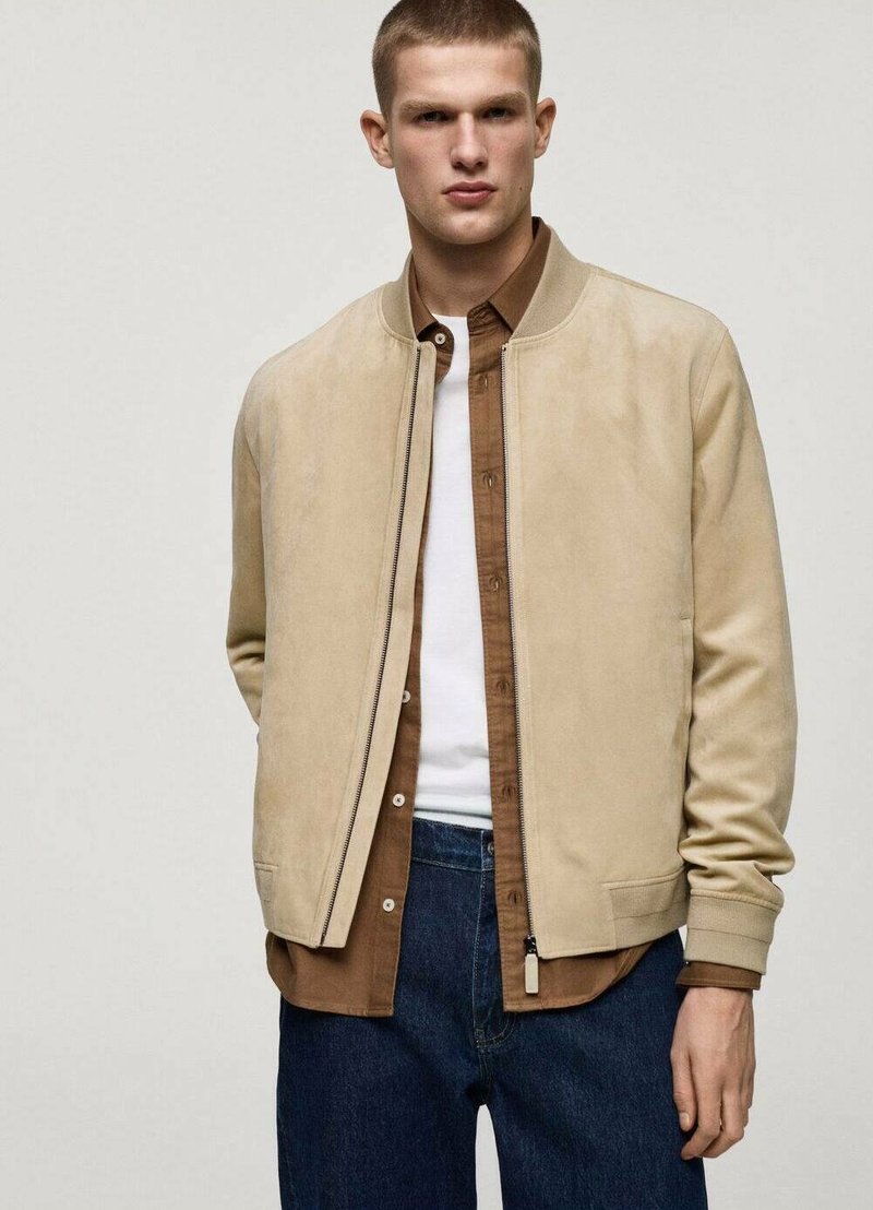 Beige bomberjakke med en glat ruskindsfinish, ribbede mansjetter og lommer, lagdelt over en brun skjorte og en hvid t-shirt.