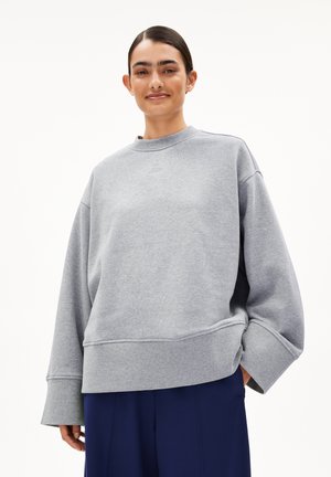 Vrouw met donker haar die een losse grijze sweater en marineblauwe wijde broek draagt, staand tegen een vlakke witte achtergrond.