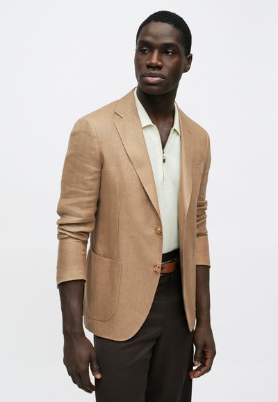 Ανδρας με σκουρόχρωμο δέρμα που φοράει μπεζ σακάκι πάνω από ένα φωτεινό πουκάμισο με φερμουάρ και σκούρο παντελόνι, κοιτάζοντας ελαφρώς προς τα δεξιά, με φόντο απλό λευκό.