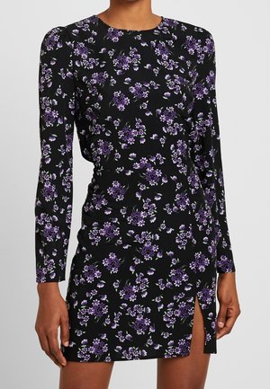 Femme portant une mini-robe noire à manches longues avec imprimé floral violet et une petite fente à l’avant de la cuisse.