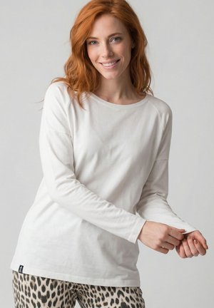 Rothaarige Frau lächelnd, trägt ein weißes Langarmshirt und Leopardenmuster-Hose, zieht ihren Ärmel vor einem schlichten Hintergrund hoch.