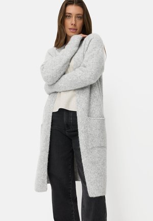 Frau trägt eine lange hellgraue flauschige Strickjacke über einem weißen Top und schwarzen hoch taillierten Hosen, steht mit verschränkten Armen nahe ihrem Gesicht.