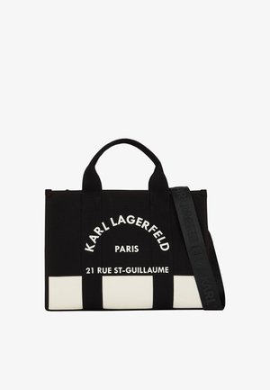 Bolsa tote de lienzo negra con texto blanco, con dos asas superiores y una correa desmontable. Incluye logo y dirección impresos en la parte delantera.