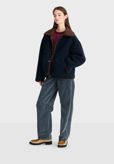 Jeune femme pose vêtue d'une veste oversize bleu marine foncé avec un col marron, un pantalon gris à jambes larges, un pull violet à motifs et des bottes beige.