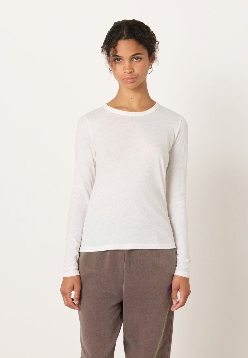 Valge pika varrukaga särk ümara kaelusega, tehtud kerge kangaga; kombineeritud lahtiste pruunide pükstega, millel on väike lilla logo.