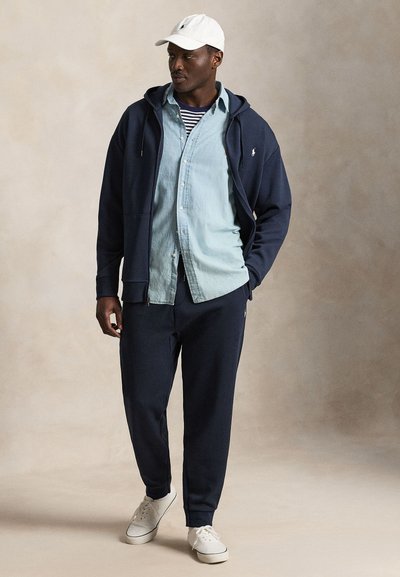 Mand iført hvid kasket, mørkeblå lynlås-hættetrøje, lyseblå cowboyskjorte over stribet top, mørkeblå bukser og hvide sneakers, står mod en ensfarvet baggrund.