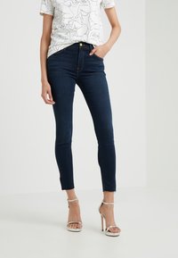 Vaqueros skinny de un índigo oscuro con una cintura alta, que presentan un cierre de botón y cremalleras en los tobillos. Combinados con una camiseta blanca que muestra un arte lineal en negro.