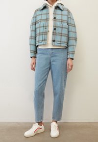 Giacca di lana a quadri blu con colletto, due tasche frontali, bottoni neri; abbinata a jeans azzurri chiari e sneakers bianche con dettagli rossi.