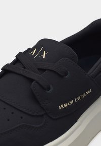 Μαύρο αθλητικό παπούτσι Armani Exchange με μαύρα κορδόνια, "A|X" στη γλώσσα και το λογότυπο "ARMANI EXCHANGE" σε χρυσό στο πλάι.