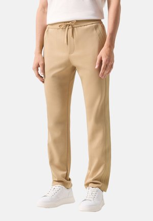 Mężczyzna w beżowych spodniach dresowych z bocznymi paskami i białych trampkach, stojący na jednolitym tle.