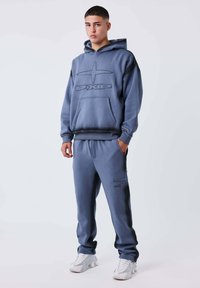 Joven de pie vistiendo una sudadera azul grisácea y un pantalón de chándal a juego con zapatillas blancas sobre un fondo blanco.
