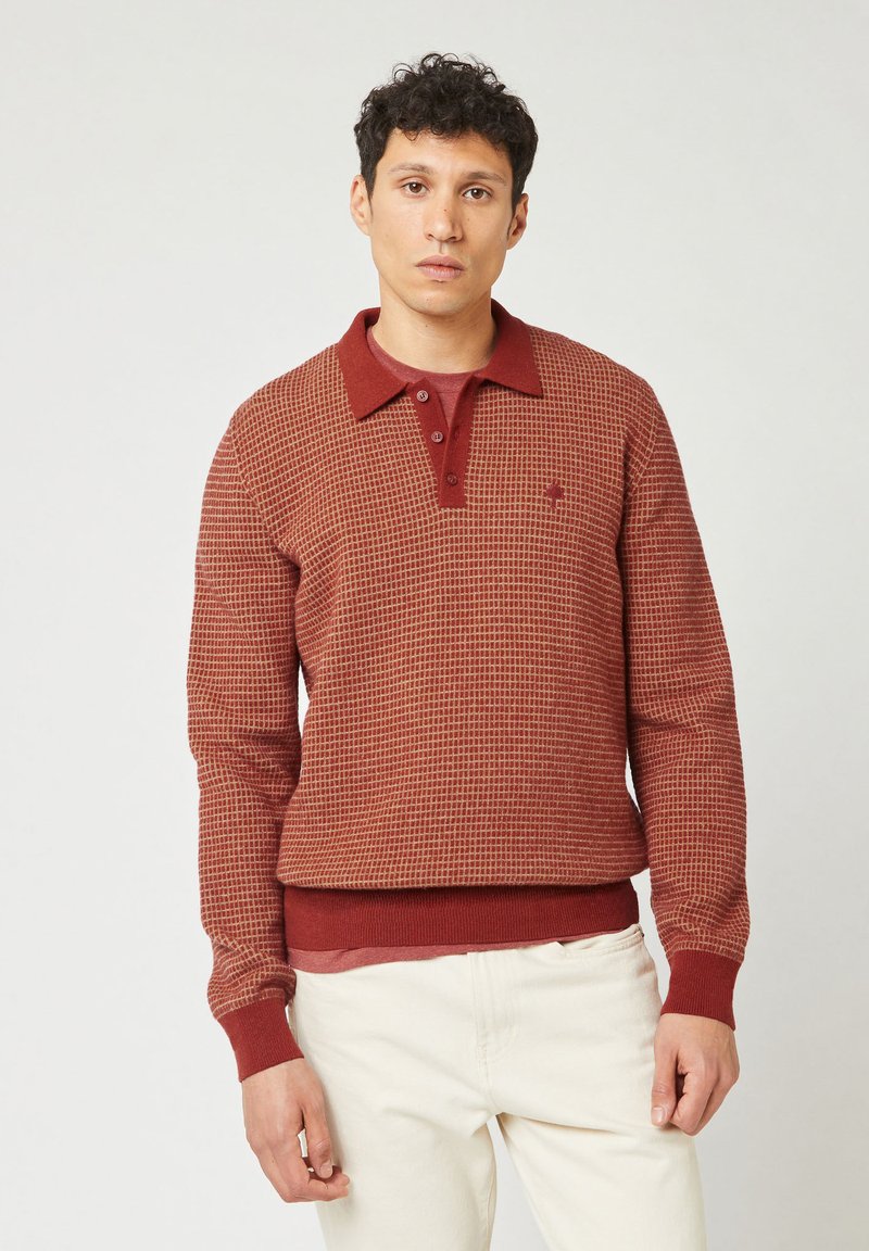 Mann in einem rot-karierten Langarmpullover mit Kragen und Knöpfen, kombiniert mit cremefarbenen Hosen, steht vor einem schlichten Hintergrund.