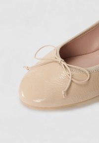 Béžové lakované baleríny s kulatou špičkou, elastickým lemem a mašlí v přední části. Hladká textura a minimalistický design.