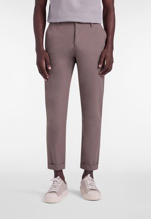 Brune smalle bukser med glat tekstur, op rullede manchetter og sidelommer, kombineret med lyse sneakers og en grå T-shirt.